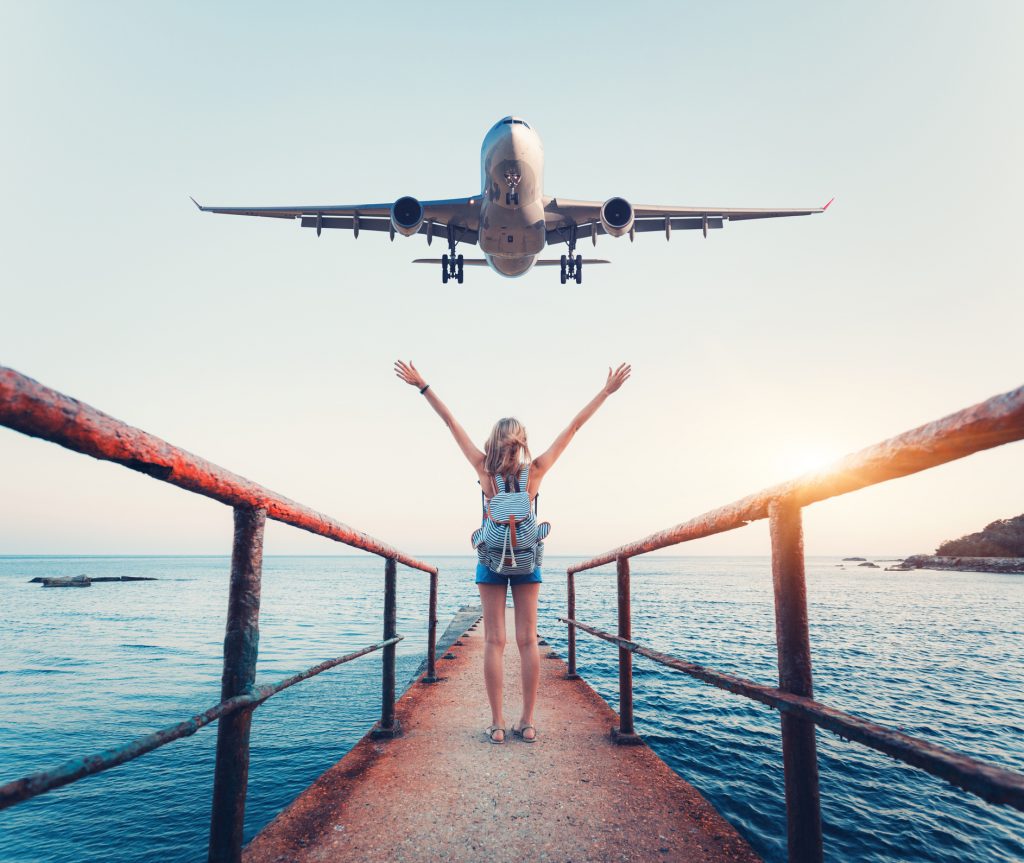 Airplane and woman at sunset. Summer landscape with girl standing on the sea pier with raised up arms and flying passenger airplane. Woman and landing commercial plane in the evening. Lifestyle