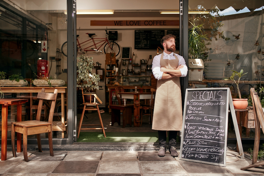 Portrait of a young man standing in front of his coffee shop. Young man with beard wearing an apron  standing with her arms crossed and looking away.