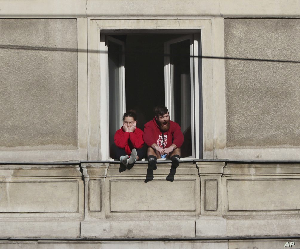 A couple bored with the government-required national isolation at home due to the spreading coronavirus, sits in the 5th floor apartment window on a sunny spring day in downtown Warsaw, Poland, Sunday, March 22, 2020.  For many people the COVID-19 coronavirus causes mild or moderate symptoms, but for some it can cause severe illness. (AP Photo/Czarek Sokolowski)