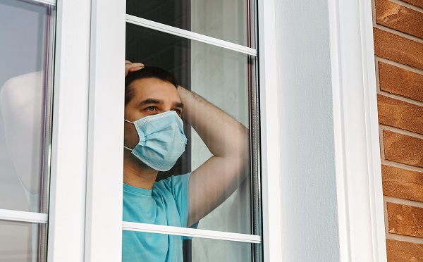 Home quarantine, self-isolation because of the Coronavirus disease, COVID-19. Man in a medical mask near the window. Boredom and depression during quarantine. Waiting for quarantine to be canceled.